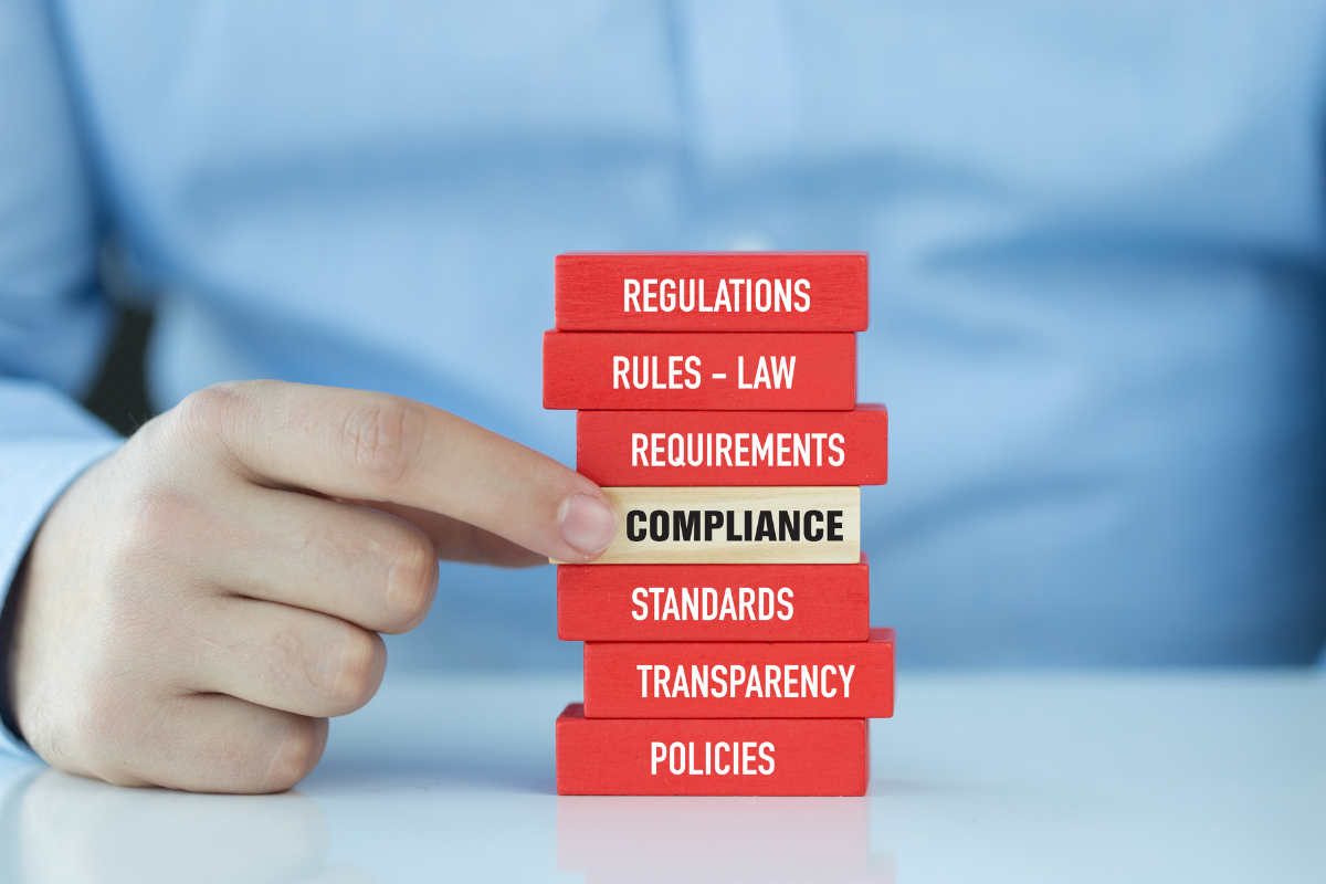 a stack of red blocks being held by a mans hand. All are red but the block that says 'Compliance' is wooden coloured to make it stand out