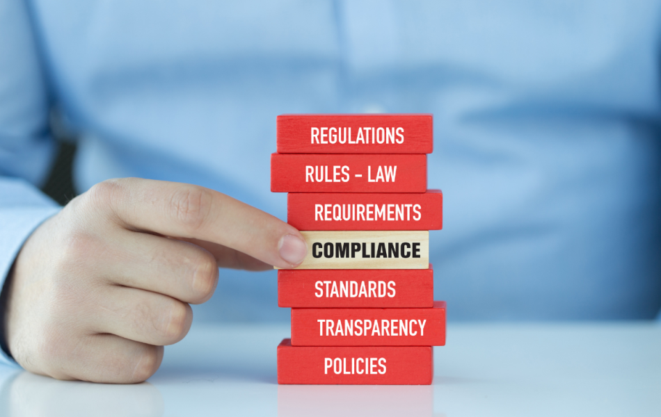 a stack of red blocks being held by a mans hand. All are red but the block that says 'Compliance' is wooden coloured to make it stand out